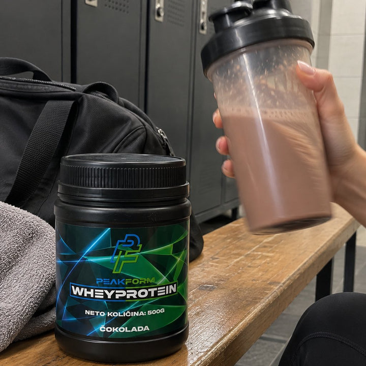 PeakForm Whey Protein čokolada 500 g na klopi v garderobi, v roki šejker s čokoladnim napitkom